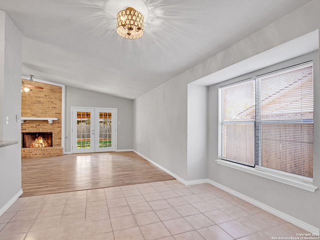 unfurnished living room with french doors, a brick fireplace, ceiling fan with notable chandelier, vaulted ceiling, and light tile patterned floors