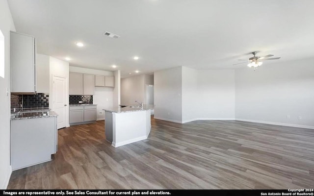 kitchen with backsplash, ceiling fan, light stone counters, an island with sink, and dark hardwood / wood-style flooring