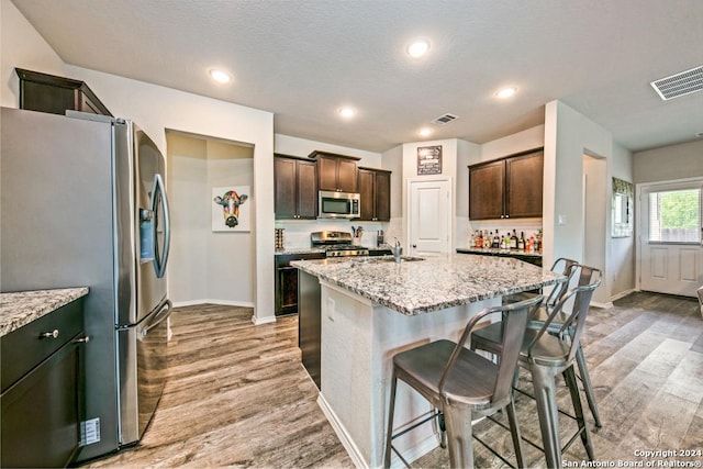 kitchen featuring light stone countertops, appliances with stainless steel finishes, light wood-type flooring, dark brown cabinets, and an island with sink