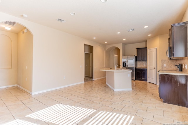 kitchen with decorative backsplash, stainless steel refrigerator with ice dispenser, dark brown cabinetry, light tile patterned floors, and an island with sink