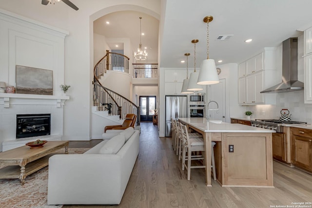 kitchen with pendant lighting, a kitchen island with sink, wall chimney exhaust hood, appliances with stainless steel finishes, and white cabinetry