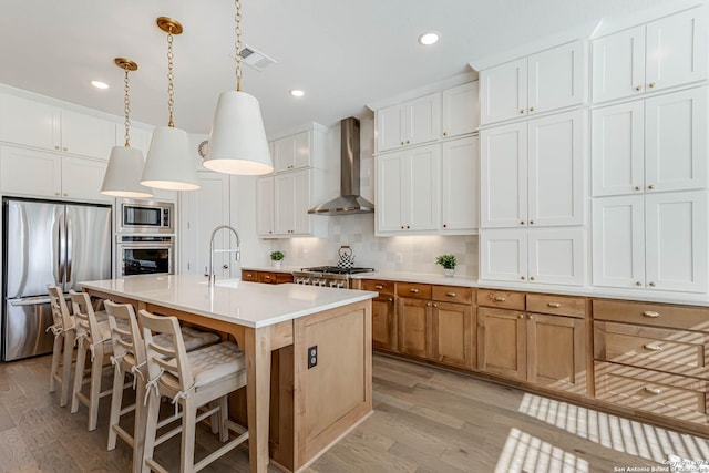 kitchen featuring a center island with sink, wall chimney exhaust hood, appliances with stainless steel finishes, decorative light fixtures, and white cabinetry