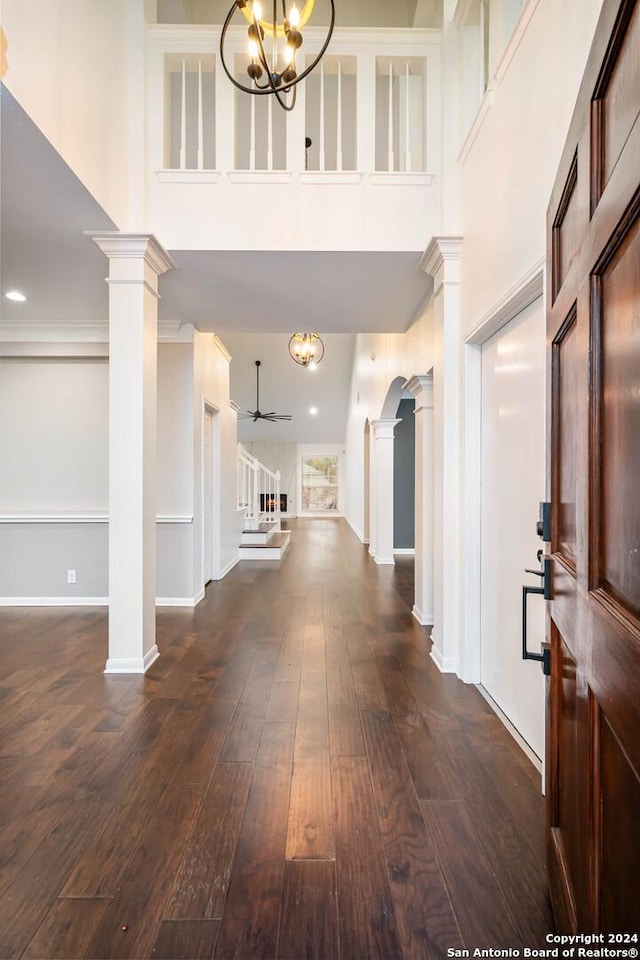 foyer featuring a towering ceiling, ceiling fan with notable chandelier, ornate columns, and dark wood-type flooring