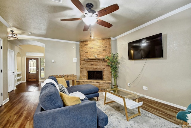 living room with a brick fireplace, a textured ceiling, ceiling fan, crown molding, and dark wood-type flooring