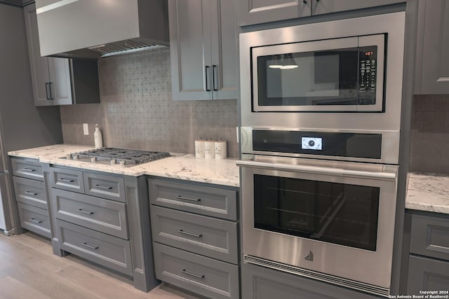 kitchen featuring light stone countertops, appliances with stainless steel finishes, backsplash, wall chimney exhaust hood, and gray cabinets