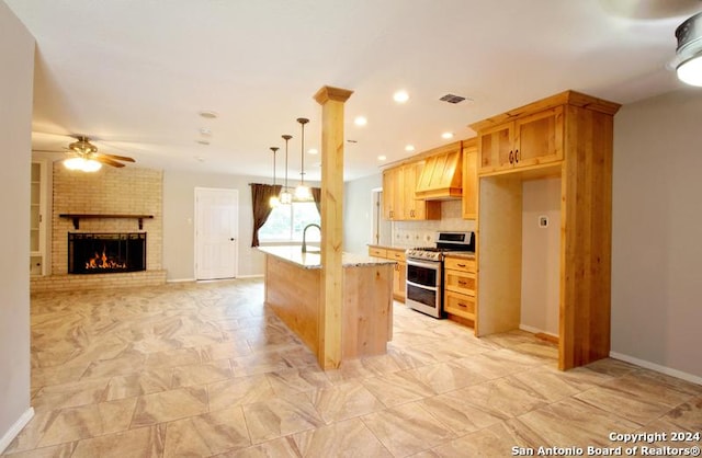 kitchen with a brick fireplace, custom range hood, ceiling fan, pendant lighting, and stainless steel range oven