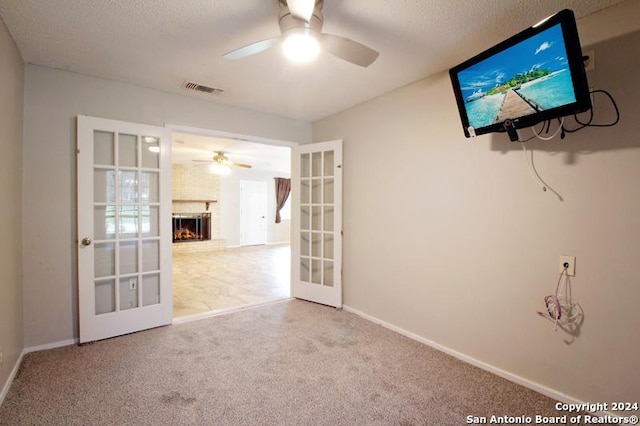 carpeted spare room featuring french doors, a textured ceiling, a brick fireplace, and ceiling fan