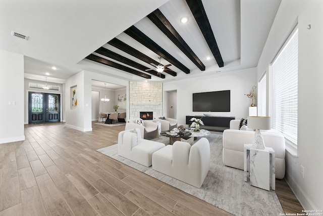 living room featuring a stone fireplace, light hardwood / wood-style flooring, beamed ceiling, and ceiling fan with notable chandelier