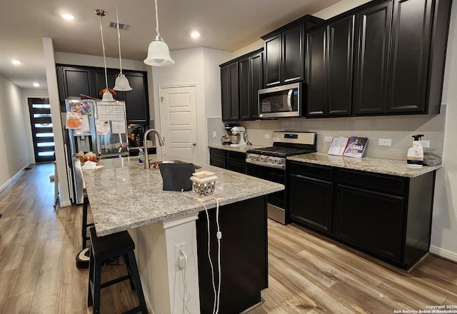 kitchen with appliances with stainless steel finishes, backsplash, pendant lighting, a center island with sink, and light hardwood / wood-style flooring