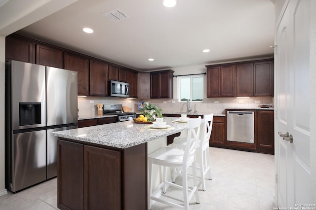 kitchen featuring light tile patterned flooring, appliances with stainless steel finishes, dark brown cabinets, a kitchen island, and light stone counters