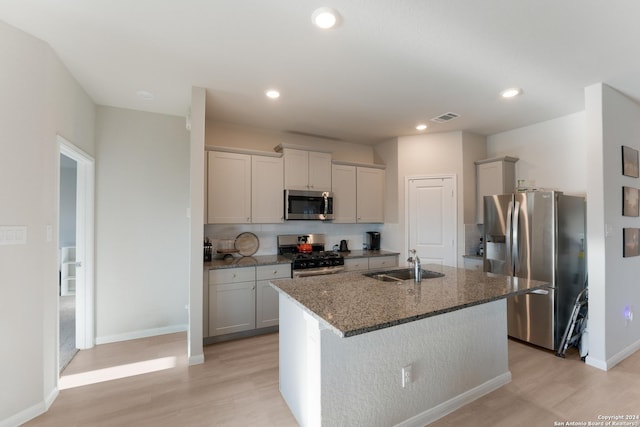 kitchen with a kitchen island with sink, dark stone counters, sink, light wood-type flooring, and appliances with stainless steel finishes