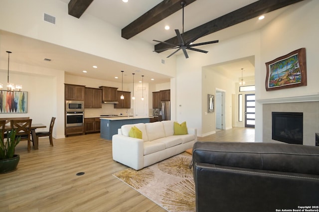 living room featuring light wood-type flooring, a towering ceiling, ceiling fan with notable chandelier, beam ceiling, and a fireplace