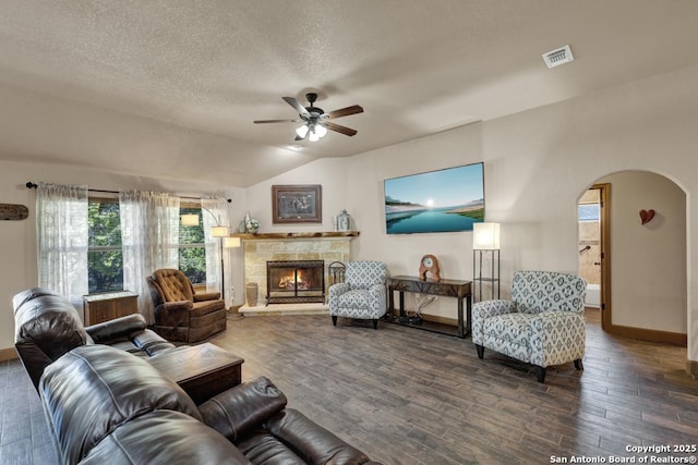 living room with a stone fireplace, ceiling fan, dark wood-type flooring, and a textured ceiling