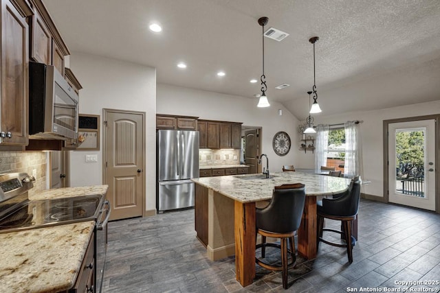 kitchen with a kitchen island with sink, hanging light fixtures, sink, decorative backsplash, and stainless steel appliances