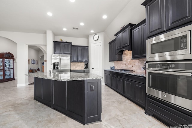 kitchen with light stone countertops, sink, stainless steel appliances, tasteful backsplash, and a center island with sink