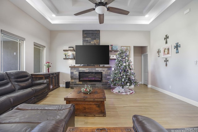 living room featuring a raised ceiling, a stone fireplace, ceiling fan, and light hardwood / wood-style floors