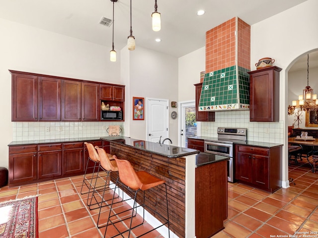 kitchen with stainless steel range with electric cooktop, a towering ceiling, an island with sink, decorative light fixtures, and black microwave