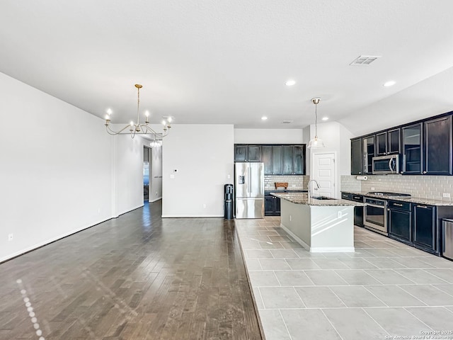 kitchen with light stone counters, an island with sink, hanging light fixtures, and stainless steel appliances