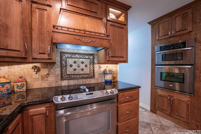 kitchen featuring tasteful backsplash, custom range hood, stainless steel appliances, light tile patterned floors, and dark stone countertops