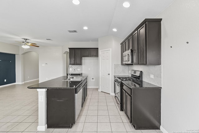 kitchen featuring appliances with stainless steel finishes, ceiling fan, sink, a center island with sink, and dark stone countertops