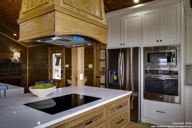 kitchen with custom exhaust hood, white cabinets, wooden walls, light stone countertops, and appliances with stainless steel finishes