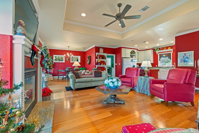 living room with ceiling fan with notable chandelier, a raised ceiling, light hardwood / wood-style flooring, a premium fireplace, and ornamental molding