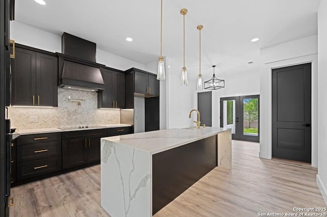 kitchen with custom exhaust hood, a center island with sink, hanging light fixtures, decorative backsplash, and light hardwood / wood-style floors