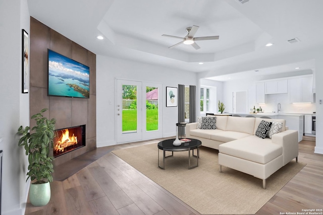 living room featuring a tile fireplace, a tray ceiling, light hardwood / wood-style flooring, and ceiling fan