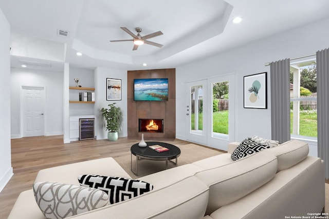 living room featuring a tile fireplace, a raised ceiling, ceiling fan, light hardwood / wood-style floors, and beverage cooler