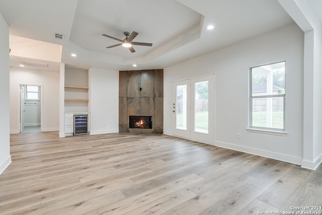 unfurnished living room featuring a large fireplace, a raised ceiling, ceiling fan, light hardwood / wood-style floors, and wine cooler