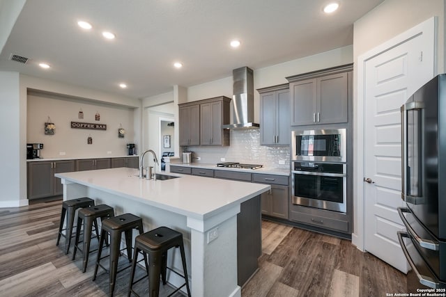 kitchen with a kitchen bar, wall chimney exhaust hood, stainless steel appliances, a kitchen island with sink, and sink