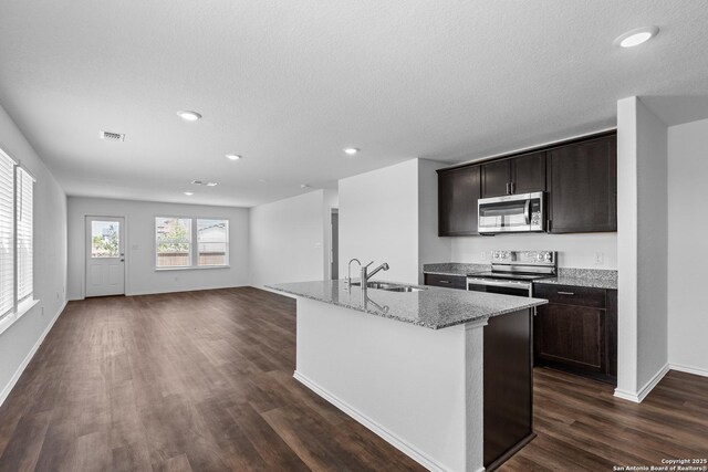 kitchen with light stone counters, dark brown cabinets, stainless steel appliances, a kitchen island with sink, and sink