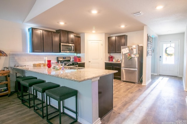 kitchen with kitchen peninsula, appliances with stainless steel finishes, light wood-type flooring, decorative backsplash, and dark brown cabinets