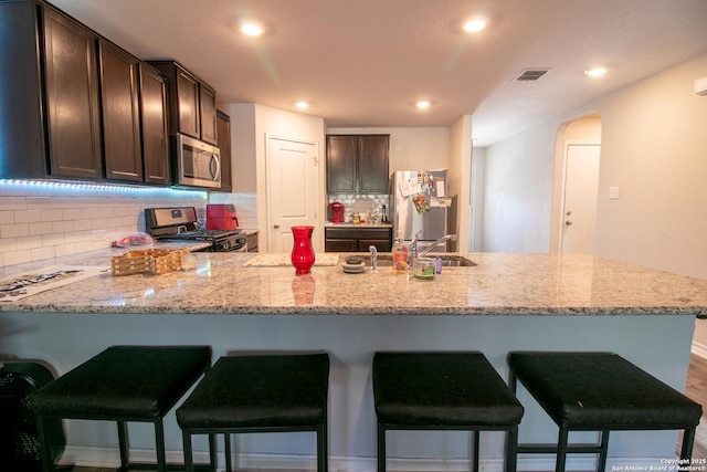 kitchen with decorative backsplash, appliances with stainless steel finishes, light stone counters, dark brown cabinets, and a breakfast bar area