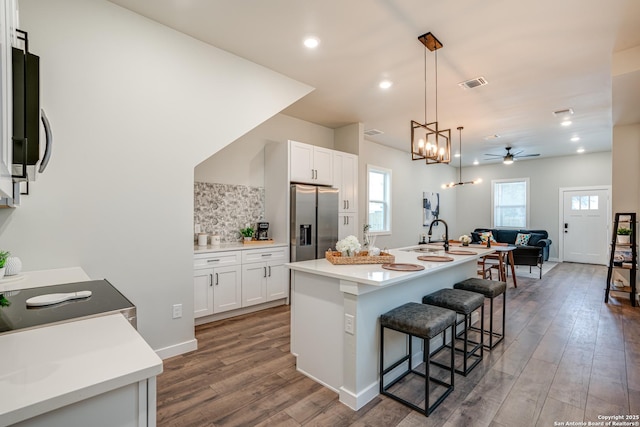 kitchen with a kitchen island with sink, ceiling fan with notable chandelier, a kitchen breakfast bar, stainless steel fridge with ice dispenser, and white cabinetry