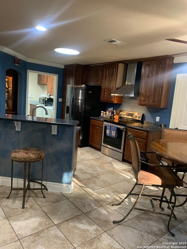 kitchen featuring stainless steel electric stove, a kitchen breakfast bar, black fridge, wall chimney range hood, and ornamental molding