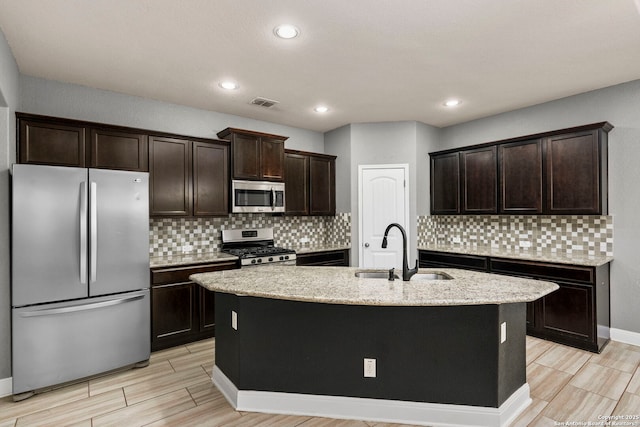 kitchen with dark brown cabinetry, sink, stainless steel appliances, light stone counters, and an island with sink