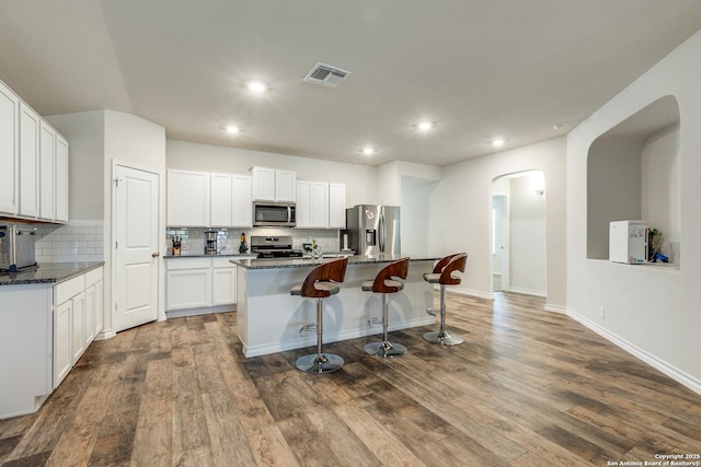 kitchen with white cabinets, stainless steel appliances, dark stone countertops, and an island with sink