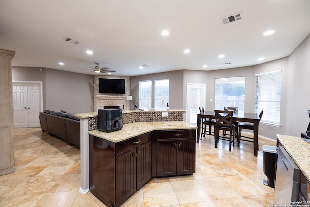 kitchen featuring a stone fireplace, ceiling fan, dark brown cabinets, and light stone countertops