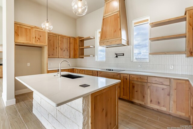 kitchen featuring custom range hood, black electric cooktop, sink, decorative light fixtures, and an island with sink