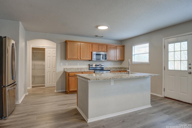 kitchen featuring sink, light stone counters, light hardwood / wood-style flooring, an island with sink, and appliances with stainless steel finishes