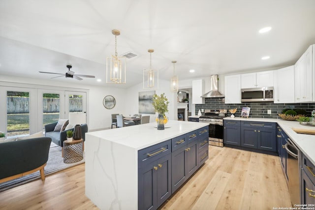 kitchen with white cabinets, wall chimney range hood, hanging light fixtures, blue cabinetry, and stainless steel appliances