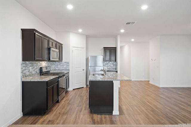 kitchen with backsplash, light stone countertops, a kitchen island with sink, and appliances with stainless steel finishes