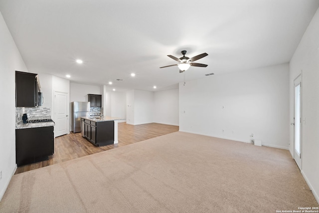 kitchen featuring decorative backsplash, stainless steel fridge, ceiling fan, an island with sink, and light hardwood / wood-style floors