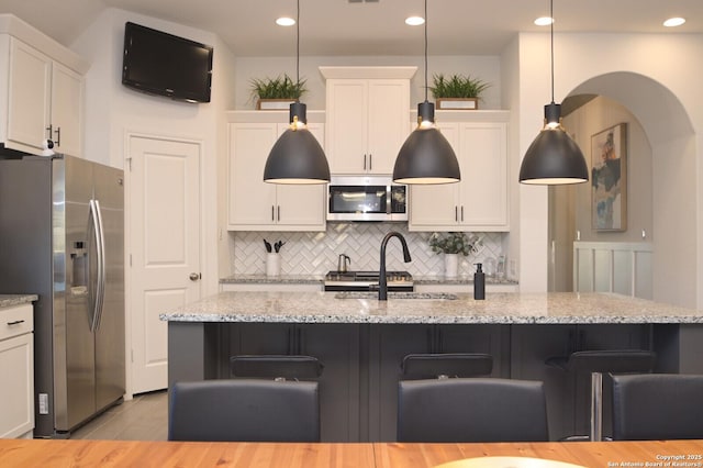 kitchen featuring decorative backsplash, light stone counters, stainless steel appliances, a center island with sink, and hanging light fixtures