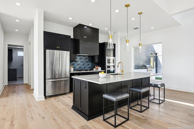 kitchen with a breakfast bar, a center island with sink, hanging light fixtures, light hardwood / wood-style flooring, and appliances with stainless steel finishes