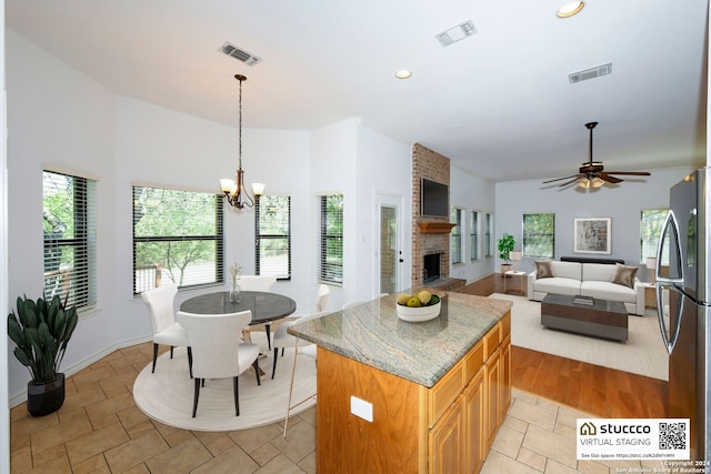 kitchen with ceiling fan with notable chandelier, a center island, a brick fireplace, stainless steel fridge, and light stone counters