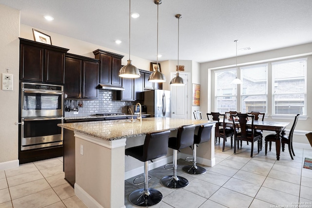 kitchen with light stone countertops, stainless steel appliances, an island with sink, decorative light fixtures, and decorative backsplash