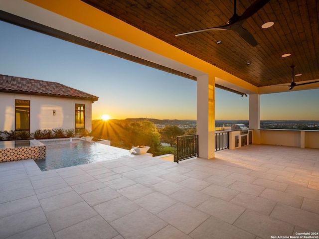 patio terrace at dusk with pool water feature, ceiling fan, area for grilling, and an outdoor kitchen