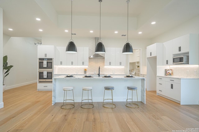 kitchen featuring white cabinetry, wall chimney exhaust hood, an island with sink, decorative light fixtures, and appliances with stainless steel finishes